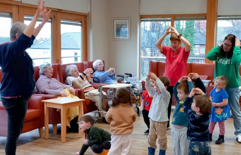 At Abbeyfield, broad smiles light up the room as older adults and young children take part in a lively group activity. Some relax on sofas, while others stand and move around with arms raised, creating a joyful and dynamic scene.