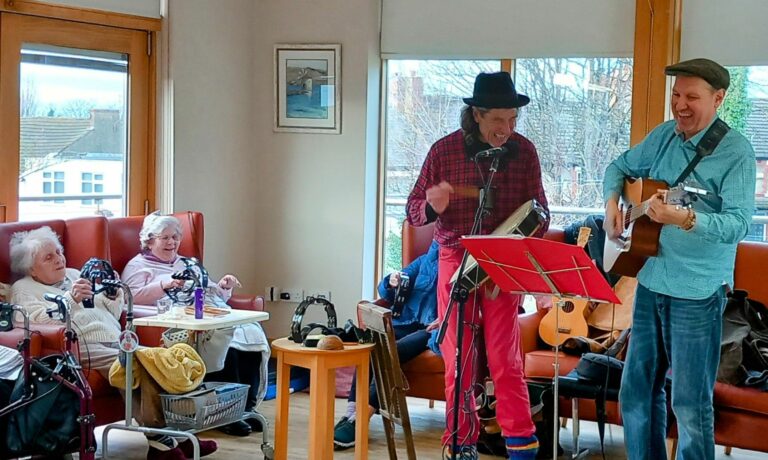 In the warm, inviting atmosphere of Abbeyfield Care Home, two musicians perform joyfully with a guitar and an accordion for seated elderly residents.