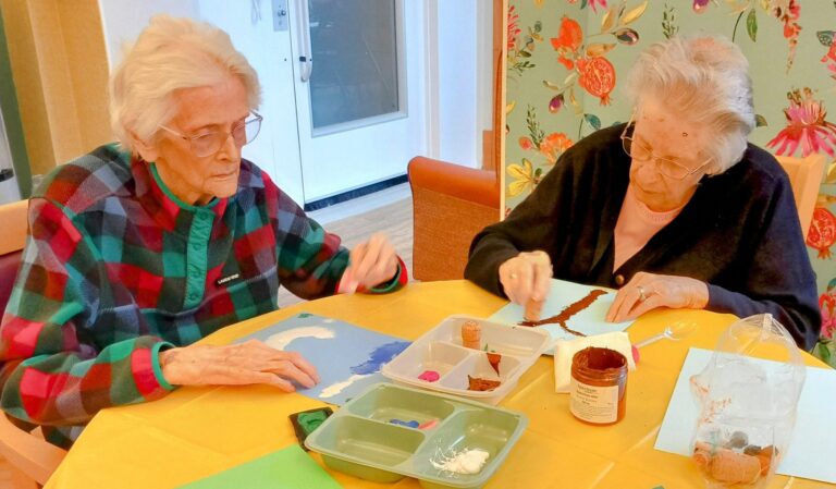 Two elderly women are enjoying a delightful crafternoon at the table, surrounded by art supplies and colourful paper, as they immerse themselves in their painting activity.