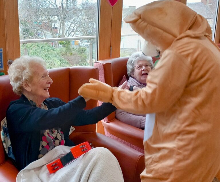 A person in an animal costume brings joy on Valentines Day at Abbeyfield Hoylake and West Kirby Society, engaging with two elderly women seated in a room with large windows overlooking the trees. The women appear amused and are smiling warmly at the lively interaction.