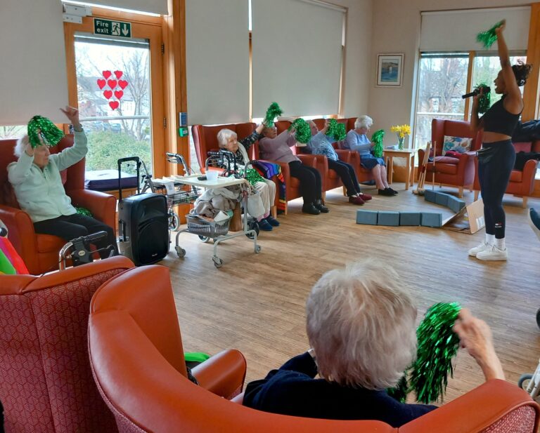 Elderly residents at Abbeyfield Hoylake and West Kirby Society enjoy a day of fun and dance, participating in a seated exercise class. With green pom-poms in hand, they are guided by an enthusiastic instructor leading the lively session.