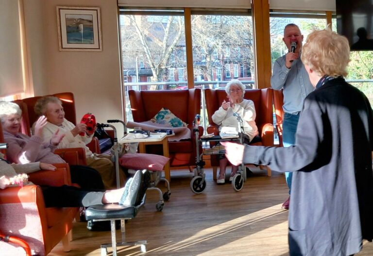 Mark Birch, known as the voice of Abbeyfield, captivates a group of seated elderly women in a sunlit room with his microphone skills. One woman in a pink jumper claps along, whilst another in black stands facing the lively gathering.