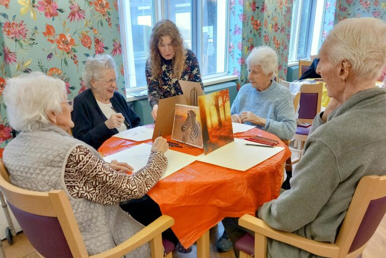 Four people gather around a table laden with art supplies, guided by an instructor. In this bright room adorned with floral wallpaper, their crafternoon unfolds into an inspiring blend of creativity and connection as they delve into drawing activities together.
