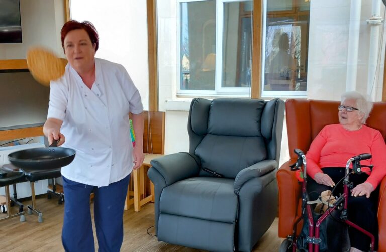 On Pancake Day, a woman in a white shirt skilfully flips a pancake in a pan, while an elderly woman with white hair and glasses observes from her chair at Abbeyfield Lear House.