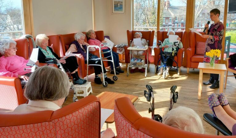 In the bright afternoon light, a group of elderly people sit in a circle of armchairs. Some use walking aids or wheelchairs as Anne stands reading poetry to them. Yellow flowers brighten the nearby table.