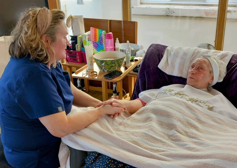 A carer in blue scrubs relaxes with an elderly person in a recliner, covered with a striped blanket, in a bright room adorned with personal items on a nearby table.