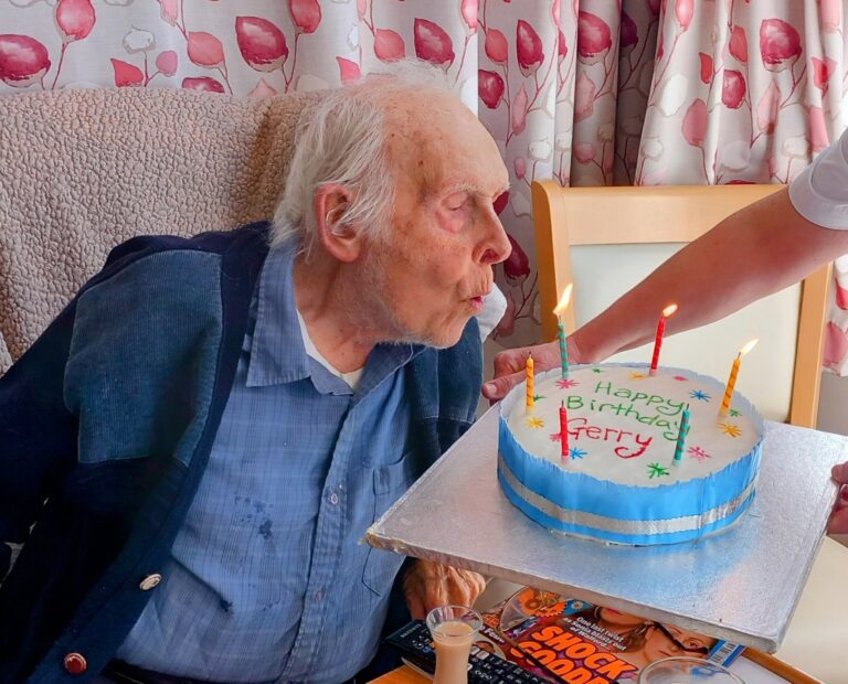 Gerry, wearing a blue jumper, smiles warmly while blowing out candles on a cake labelled Happy Birthday, as a hand holds the sweet surprise.