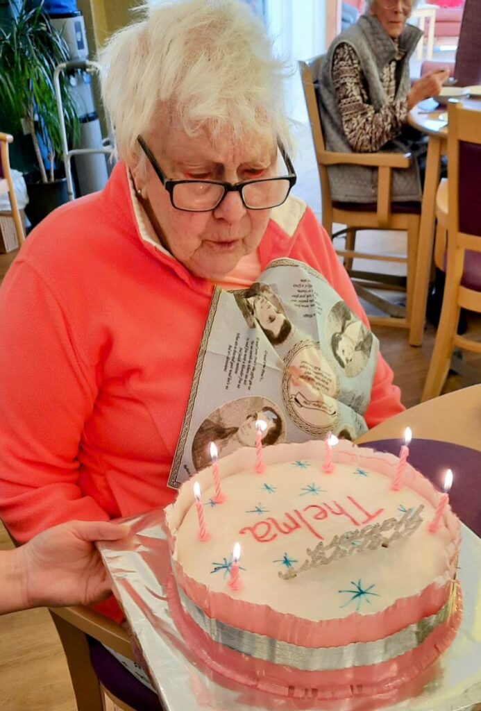An elderly woman with white hair and glasses enjoys some birthday fun, holding a pink cake with candles and the name Thelma on it. Another person is seated in the background. In British English: An elderly woman with white hair and glasses is enjoying some birthday fun, holding a pink cake adorned with candles and the name Thelma. Another person is seated in the background.