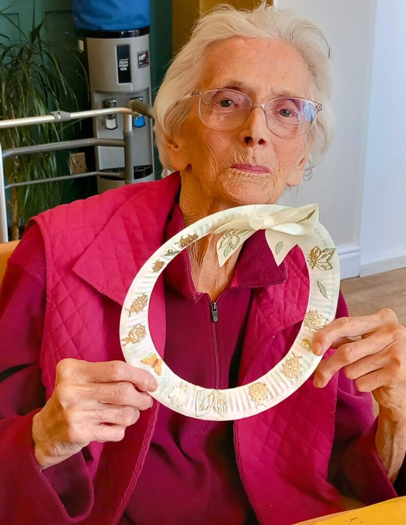 Celia, an elderly woman in a maroon jacket, holds a decorated paper wreath with a bow during Craft Afternoon while sitting at a table indoors.