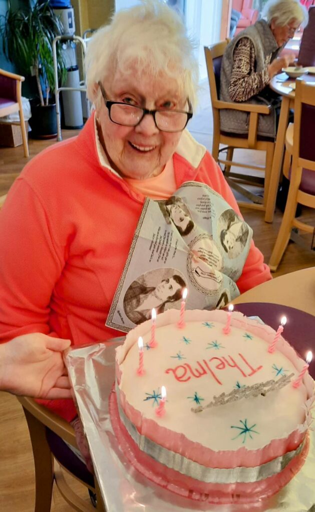 An elderly woman wearing glasses smiles as she holds a birthday cake with pink candles and the name Thelma written on it, enjoying some birthday fun while also holding a card as she sits indoors.
