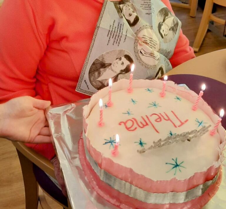An elderly woman wearing glasses smiles as she holds a birthday cake with pink candles and the name Thelma written on it, enjoying some birthday fun while also holding a card as she sits indoors.