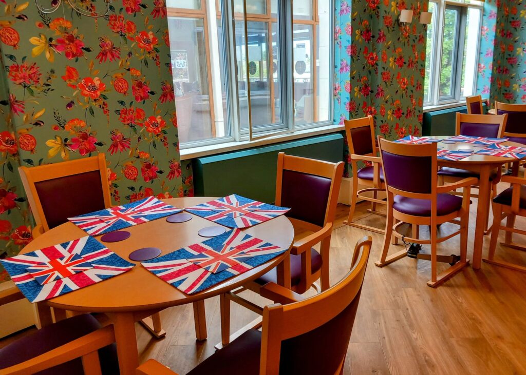 A dining area with wooden tables and chairs, Union Jack placemats, floral wallpaper, and large windows—perfect for a memorable VE Day celebration or commemorating the 80th anniversary.