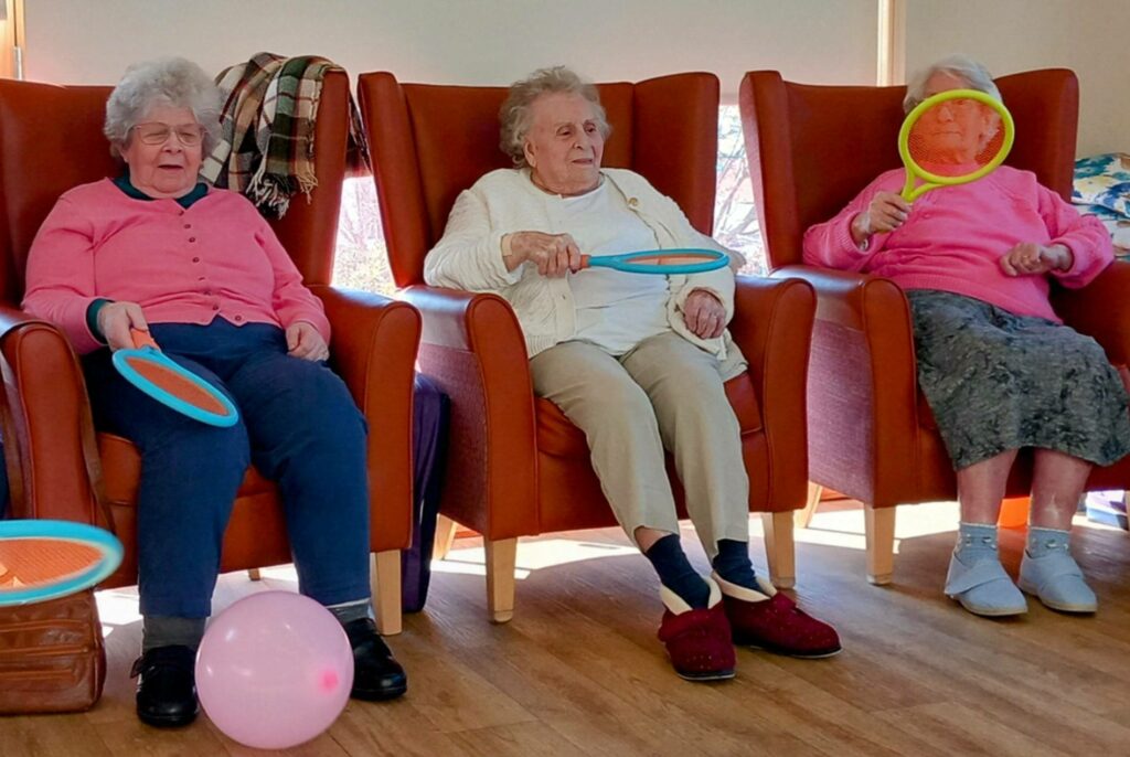 Three elderly women enjoy activities with Carla, seated in armchairs holding colourful racquets, while a pink balloon rests on the floor. One woman playfully holds her racquet in front of her face.