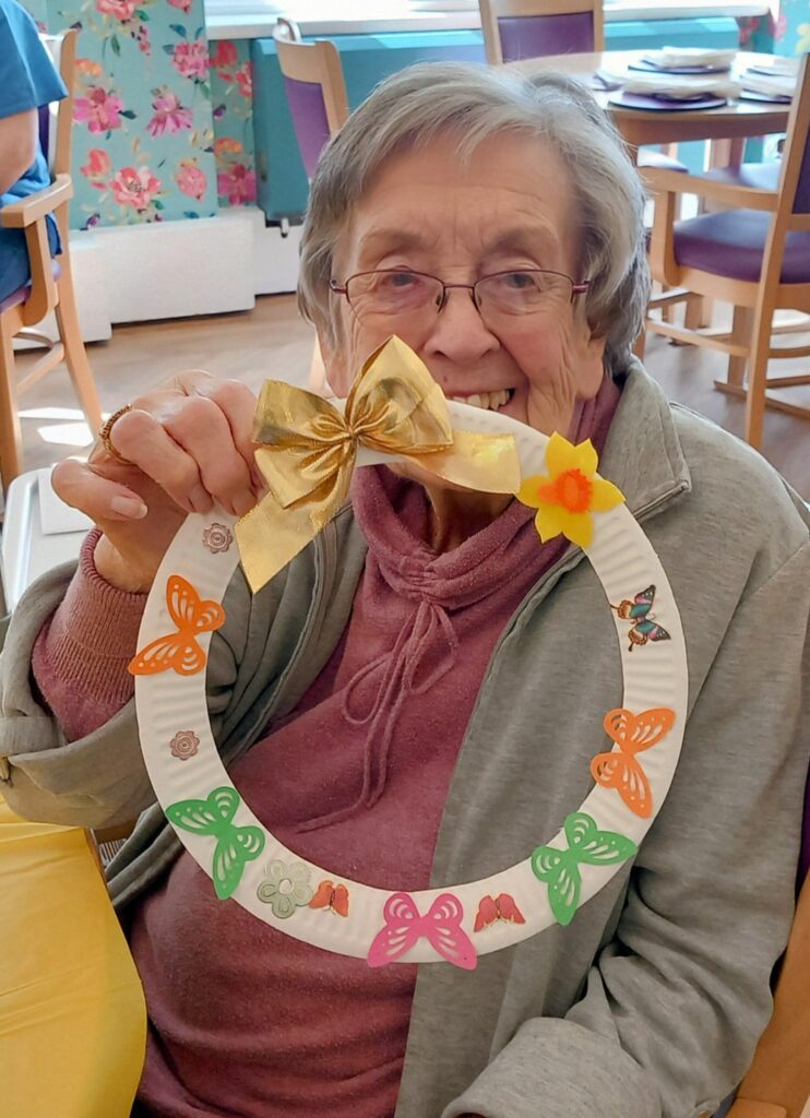 Celia enjoys a fabulous afternoon of crafts, smiling as she holds up her handmade wreath adorned with colourful butterfly cut-outs, a yellow bow, and a flower.