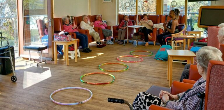A group of elderly people sit in a bright room with chairs arranged in a circle; several hula hoops are placed on the floor in the centre for activities with Carla.