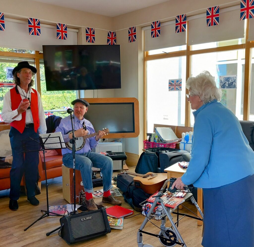 Three elderly people are in a decorated room with Union Jack flags; two men perform music while a woman with a walking frame listens, celebrating the 80th Anniversary of VE Day.