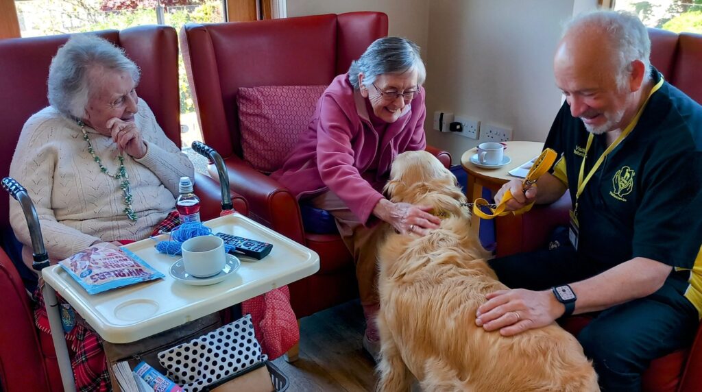 Two elderly women sit in armchairs, stroking a golden retriever during Tim’s visit, while a man in uniform looks on. Tea cups, a saucer, magazines and a TV remote rest on the table nearby.