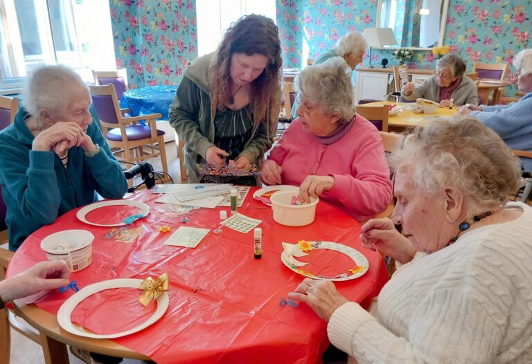 During Craft Afternoon, Celia and a group of elderly women sit around a table making crafts out of paper plates, assisted by a younger woman in a brightly lit room.