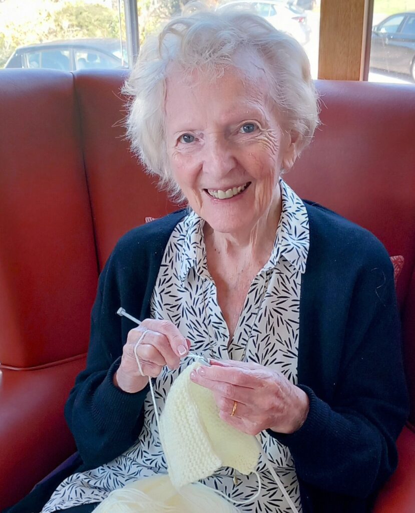 An older woman sits on a red chair, smiling as she crochets with yellow wool and holds a crochet hook at a Knit and Natter event.