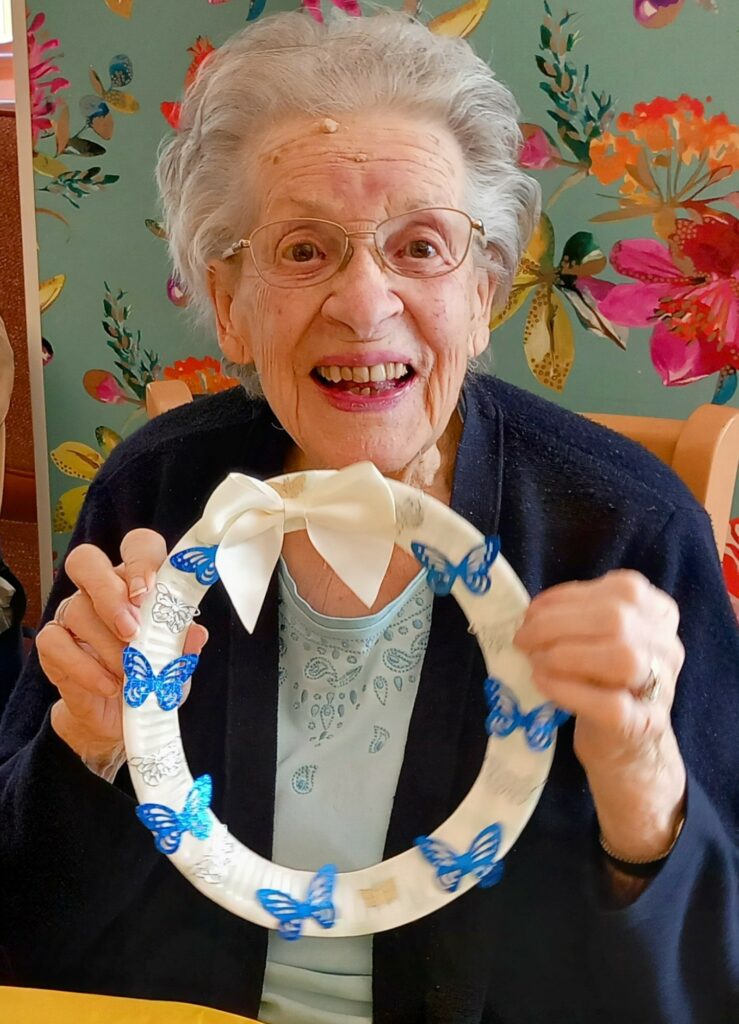 Celia enjoys a fabulous craft afternoon, smiling as she holds a white decorative wreath adorned with blue butterflies and a white bow, seated in front of floral wallpaper.