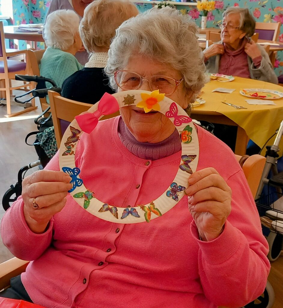 Celia, wearing a pink jumper, proudly holds up a decorated craft ring with butterfly stickers and bows during a Craft Afternoon in the communal dining area.