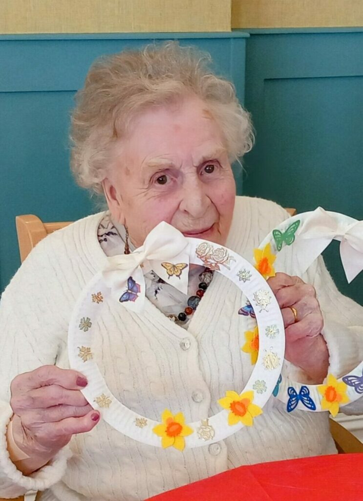 Celia and another woman display decorated wreath crafts with flowers and butterflies while seated at a table during a Craft Afternoon.