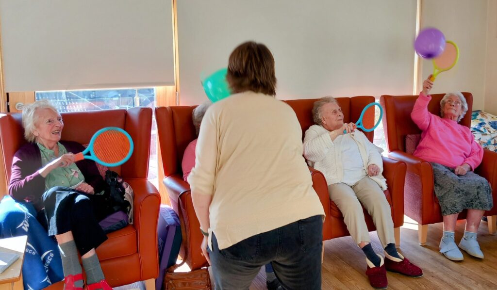 Four elderly women sit in armchairs using plastic racquets to hit balloons, while another person—Carla—stands facing them, facilitating engaging activities with them.