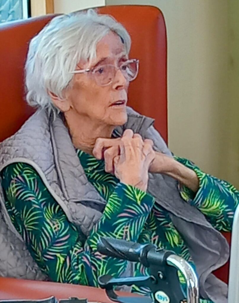 Elderly woman with white hair and glasses sits in an orange chair, wearing a patterned green top and grey waistcoat, hands clasped in front of her, with a mobility aid nearby on the VE Day anniversary.