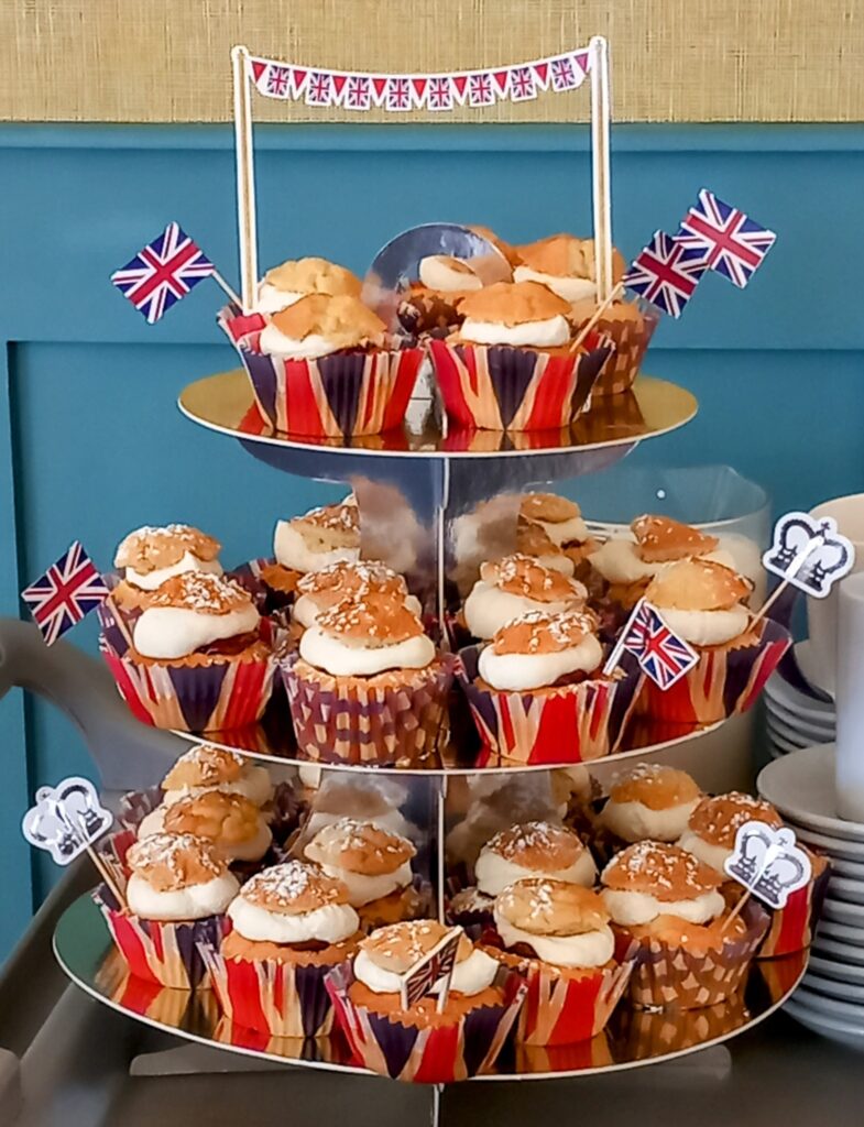 Three-tier cupcake stand with cream-filled cupcakes, decorated for the VE Day 80th Anniversary with Union Jack flags and British-themed toppers, next to a stack of white plates.