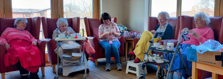 Five women—four elderly and one middle-aged—sit in a sunlit room as part of a Knit and Natter group, some engaged in knitting or crocheting, with drinks and a walking frame nearby.