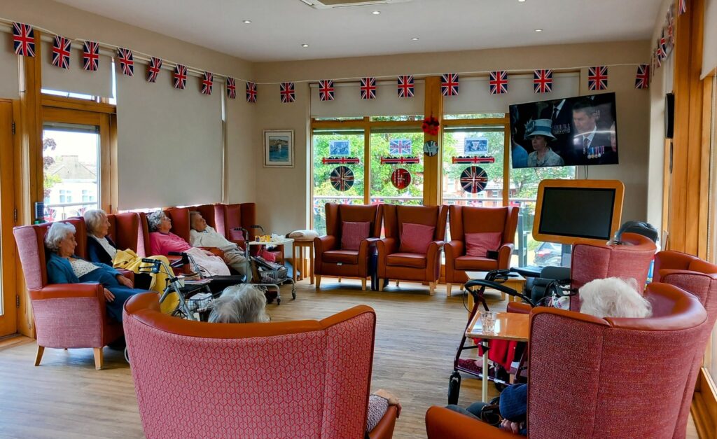 A group of elderly people sit in a lounge decorated with British flags, watching a television broadcast of Queen Elizabeth II to mark the 80th anniversary of VE Day.