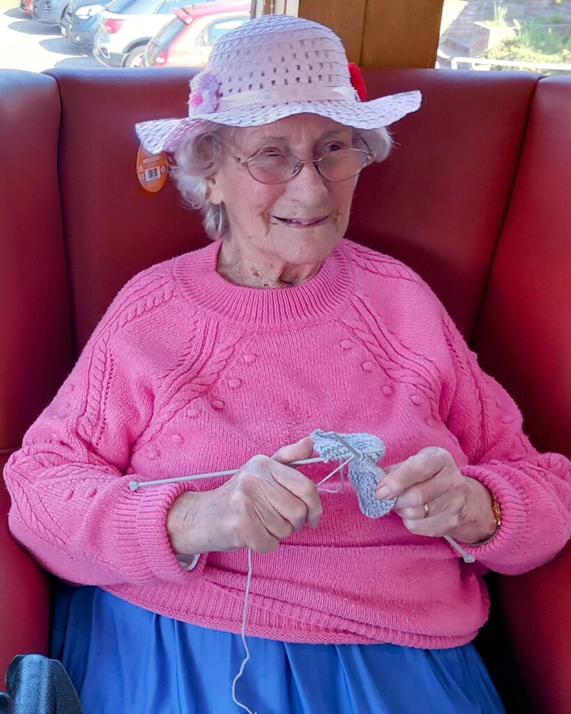 An elderly woman wearing a pink jumper and hat sits on a red chair, smiling as she knits with grey wool—enjoying an April afternoon of knitting.