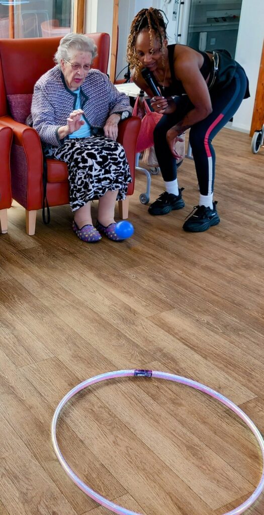 An elderly woman sits in a red chair holding a ball, next to Carla, who has a microphone. A hula hoop lies on the floor in front of them during their activities with Carla.