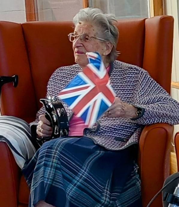 Elderly woman seated in an orange armchair, holding a tambourine and waving a Union Jack flag, dressed in a patterned jumper and checked skirt, celebrating the 80th anniversary of VE Day.