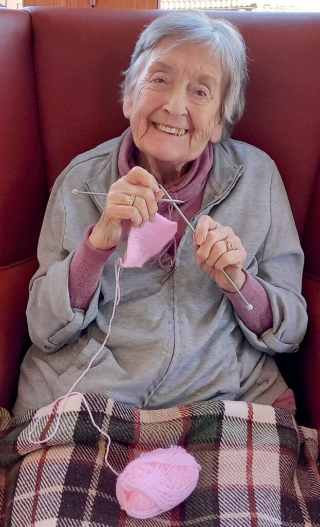 An elderly woman sits in an armchair, smiling as she knits with pink wool during a Knit and Natter. A checked blanket covers her lap and the ball of wool rests on the blanket.
