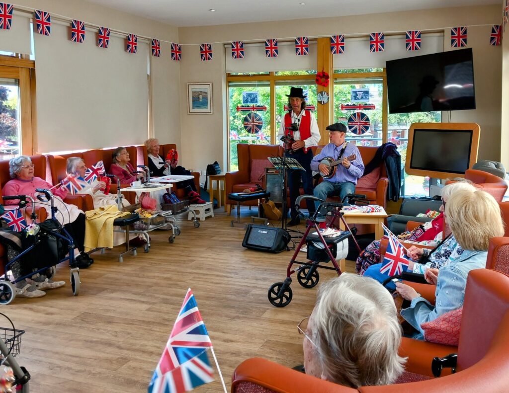 Elderly people sit in a decorated communal lounge adorned with Union Jack flags, watching two musicians perform on the 80th anniversary of VE Day; wheelchairs and walking frames are visible.