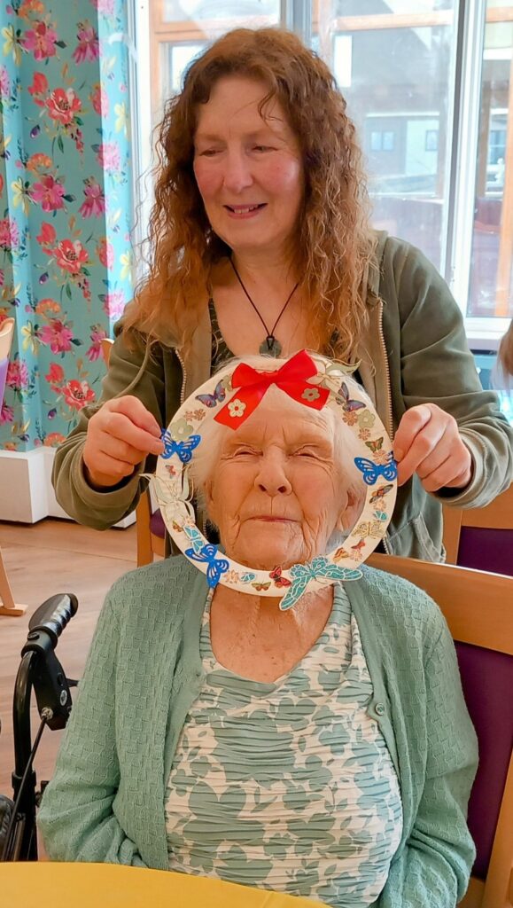 During a Craft Afternoon, Celia holds a decorated paper wreath around the face of an elderly woman seated in front of her; both are smiling in a brightly lit room.
