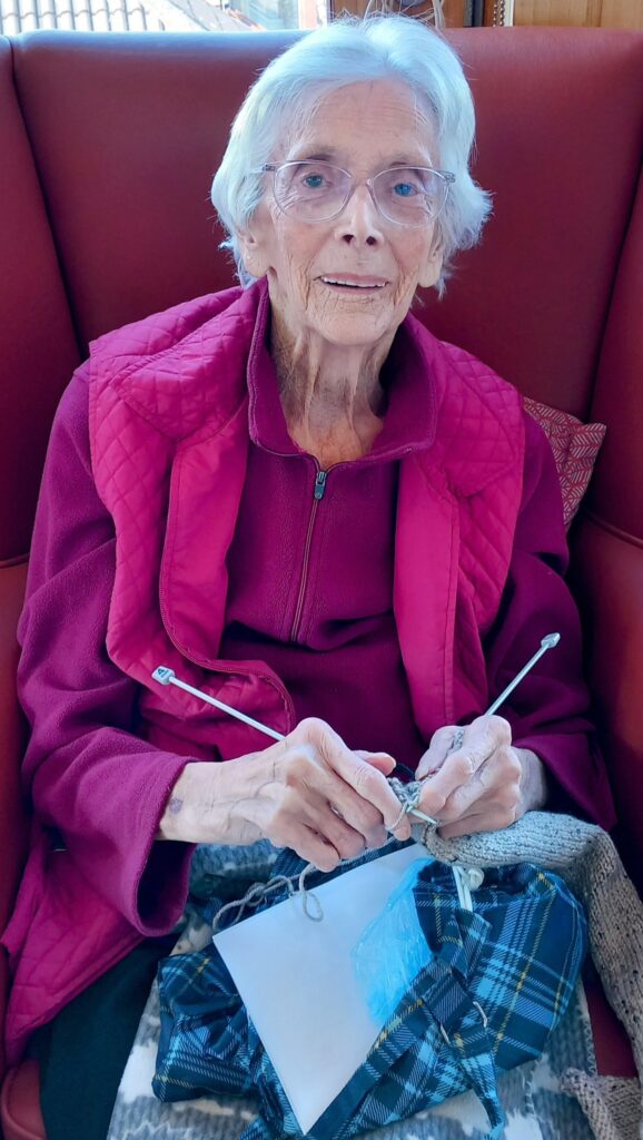 An elderly woman with white hair and glasses sits on a red chair, wearing a magenta jacket, knitting with needles and wool—perhaps at her favourite Knit and Natter group.