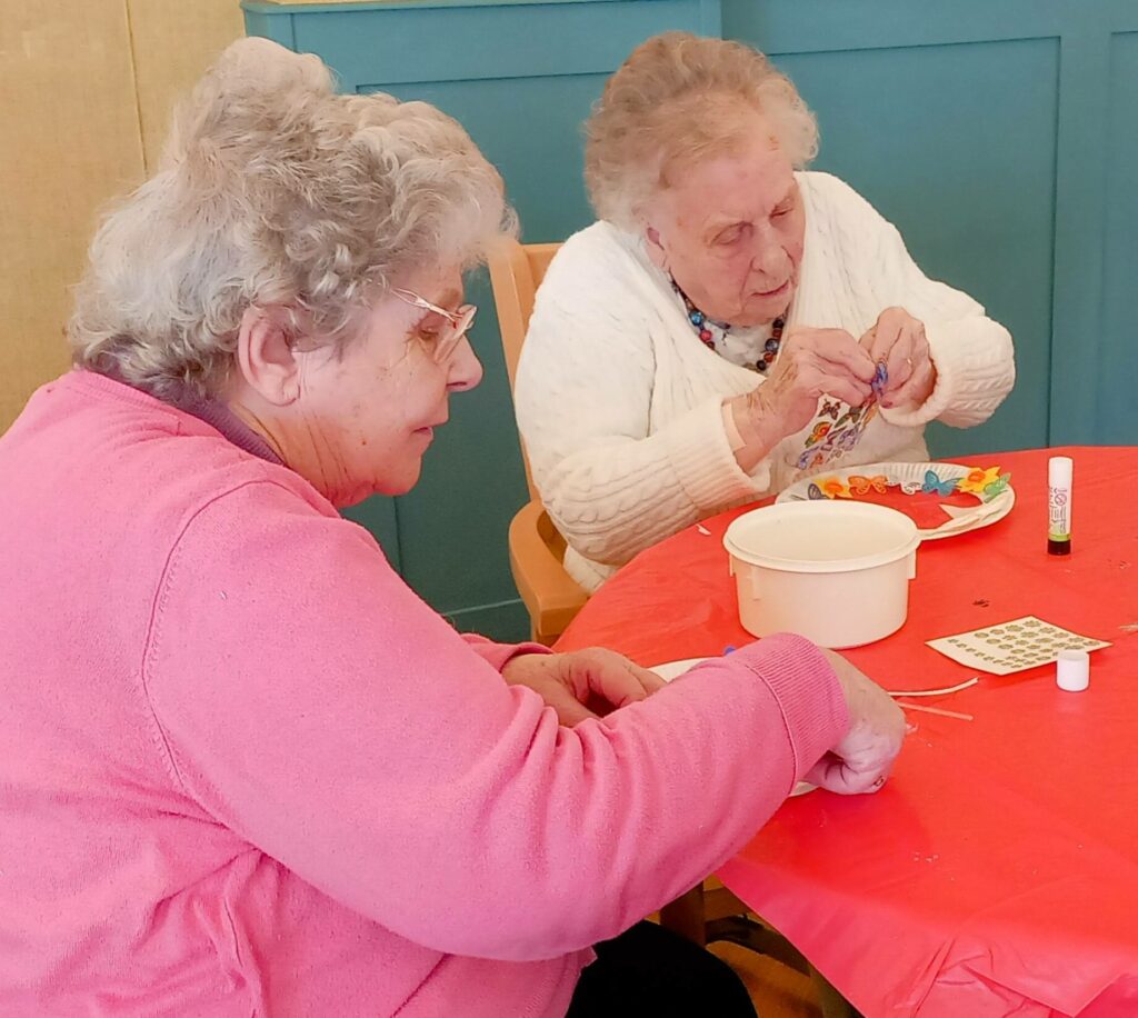 Celia and a friend sit at a red table, engrossed in their afternoon of crafts, working with small items, glue, and a container in front of them.