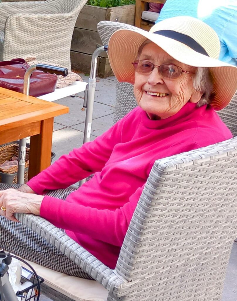 Elderly woman wearing a hat and pink jumper sits and smiles in a wicker chair outdoors, enjoying good company and the sunshine, with a walking frame and table nearby.