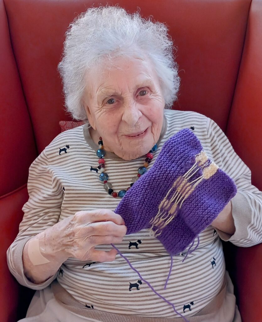 An elderly woman with white hair sits in a red armchair, smiling and holding a piece of purple knitting—ready for April Knit, the cosy knitting event. She is wearing a stripy top with dog patterns and a beaded necklace.
