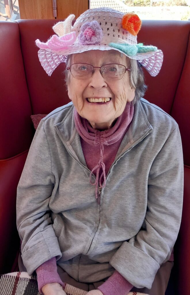 An elderly woman wearing glasses and a colourful, flowered hat sits smiling on a red chair, dressed in a grey jacket and purple scarf, ready for the Knit and Natter knitting event this April.
