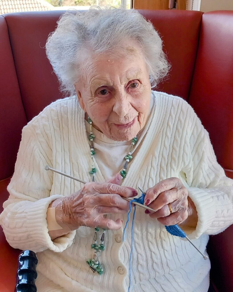 Elderly woman with white hair and a beaded necklace sits on a red chair, knitting with blue wool and silver needles at the April Knit and Natter event.