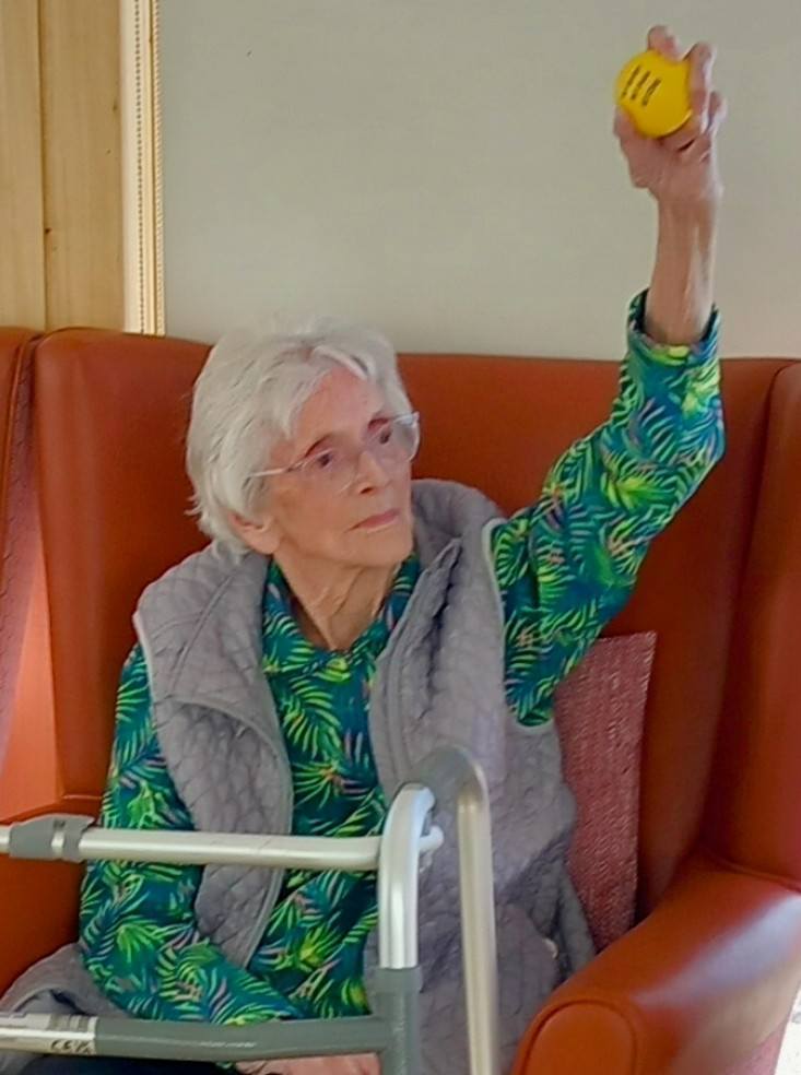 An elderly woman sits in an armchair, holding up a yellow ball in her right hand during Carla activities. A walking frame is positioned in front of her.