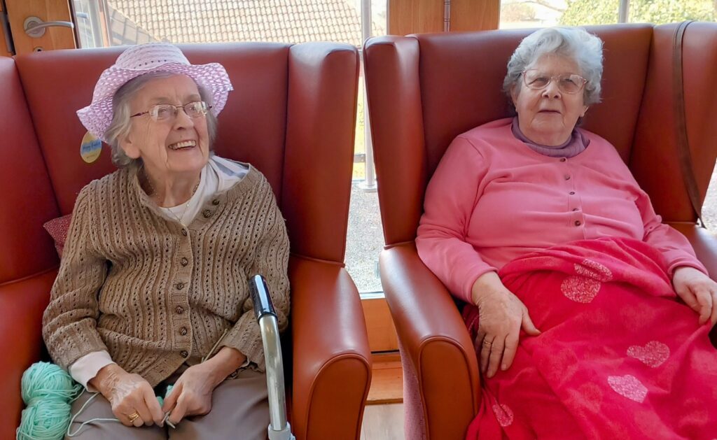 Two elderly women sit in armchairs at a Knit and Natter; one wears a sunhat and smiles with wool in her lap, while the other wears glasses, a pink jumper, and covers herself with a red blanket decorated with hearts.
