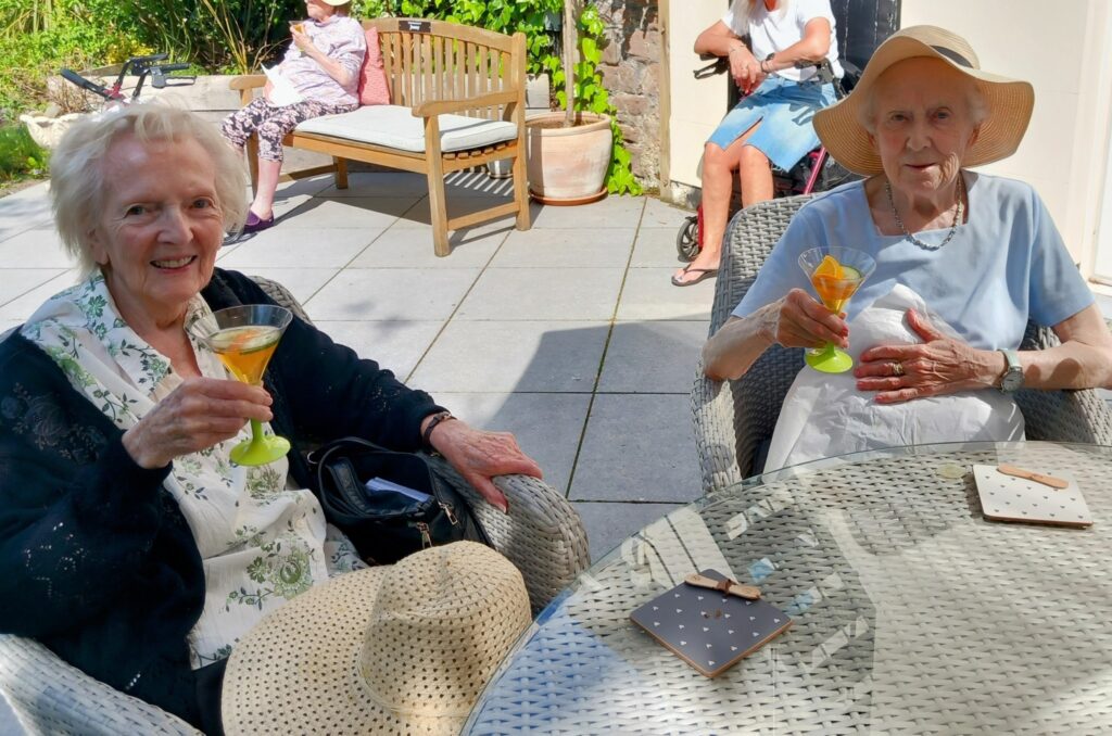 Two elderly women are seated outdoors at a glass table, each enjoying a Pimm’s. Good company and garden furniture surround them on this sunny day.