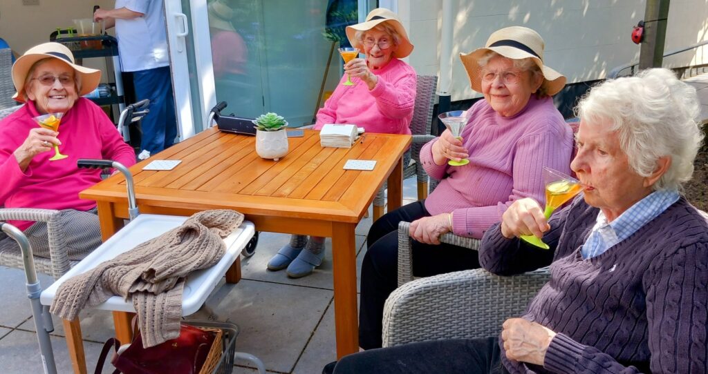 Four elderly women wearing hats are seated around a wooden outdoor table in good company, each holding a drink in a martini glass, with another person standing in the background.