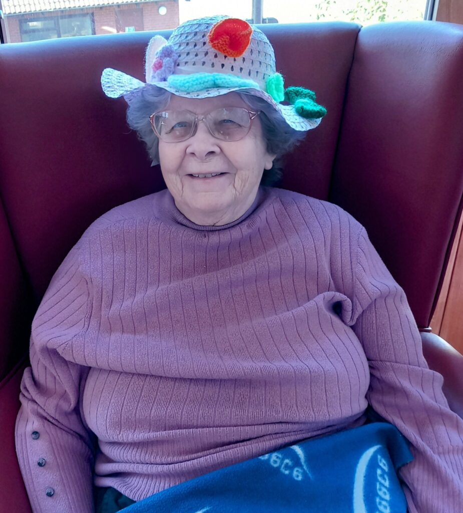 Elderly woman in a purple jumper and colourful crocheted hat sits smiling in a red chair, blanket across her lap, enjoying April knitting with her Knit and Natter group.
