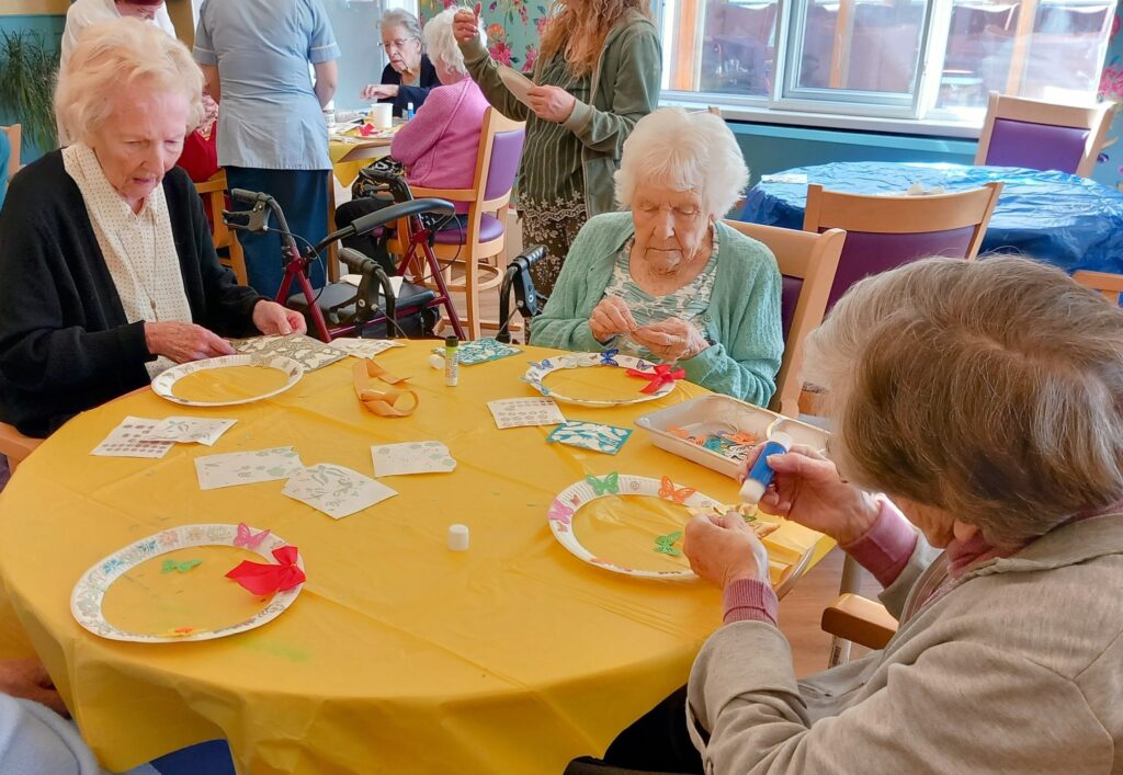 Three elderly women sit at a table covered with a yellow cloth, enjoying a marvellous afternoon of crafting with paper, glue, and decorations in a communal setting. Celia smiles as they create together.