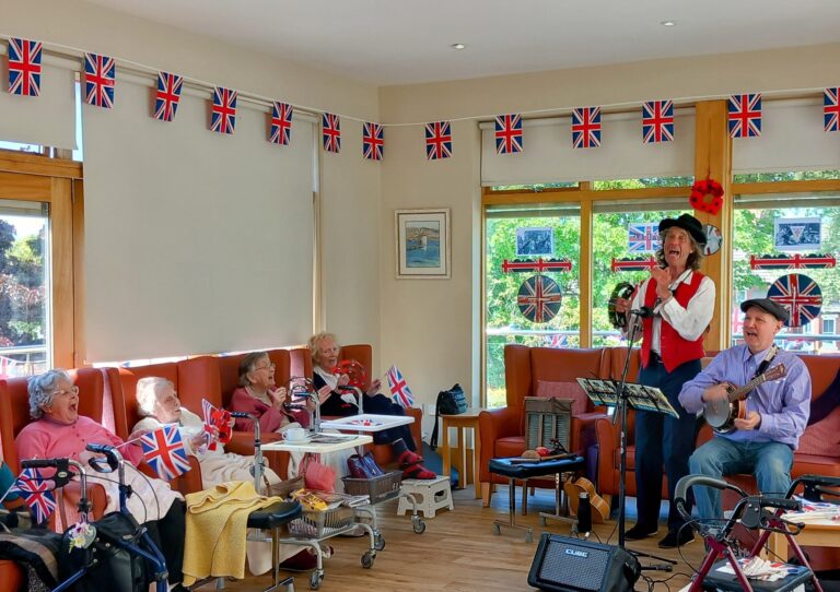 A small band performs for elderly people seated in a care home lounge decorated with numerous Union Jacks during an 80th Anniversary VE Day celebration.
